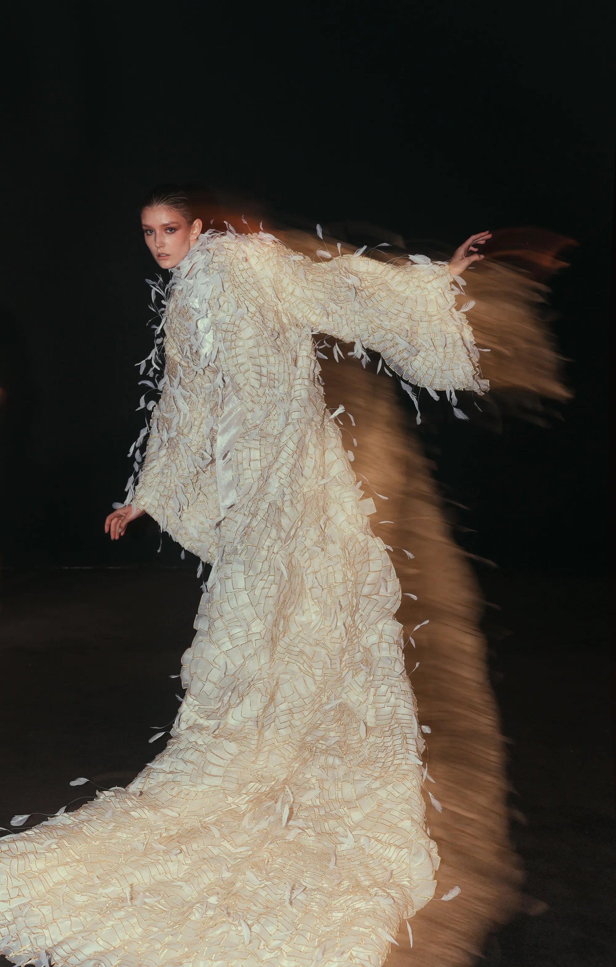 Un mannequin en robe à plumes ivoire tourne en mouvement sur un podium sombre