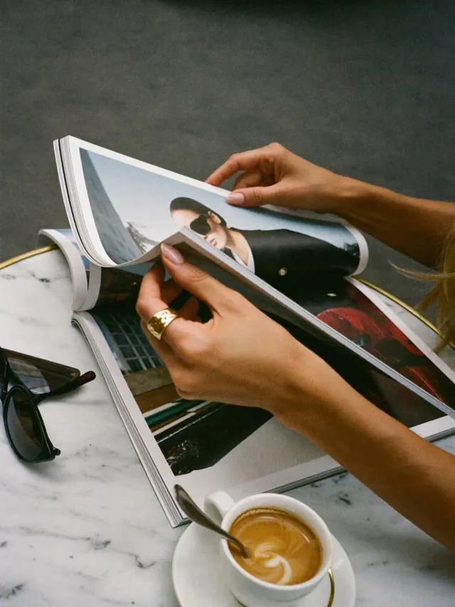 Des mains tournent les pages d’un magazine de mode à côté d’un cappuccino sur une table en marbre