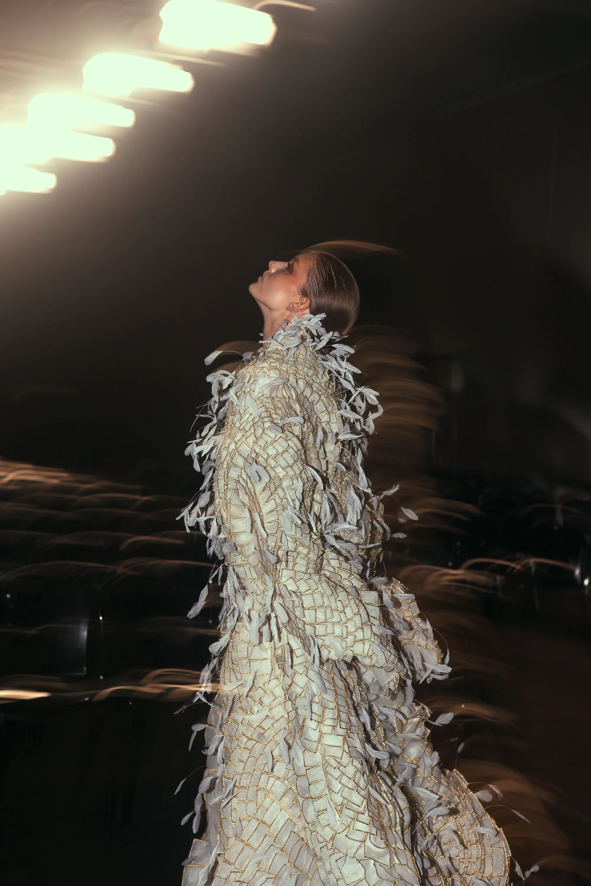 Un mannequin en manteau couture à plumes, le regard levé sous les projecteurs